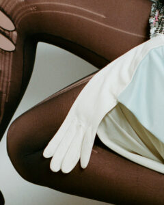 A pair of elegant white gloves rest gracefully across a surface. The background features dark, textured fabric with visible lines and seams. The composition is juxtaposed with a pale, soft garment, possibly a dress, adding a touch of sophistication. The overall scene suggests a blend of fashion elements, highlighting contrasting textures and colors in a stylish manner.