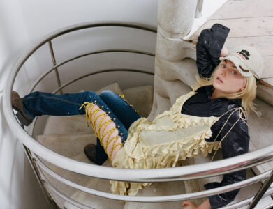 A person reclines on a spiral staircase, wearing a casual ensemble combining denim, a flowing, ruffled overlay, and a cap. The setting suggests a modern, industrial design with smooth metallic railings and light stone steps. The person's gaze is relaxed, adding a laid-back vibe. The composition highlights contrasting textures—soft fabric against hard surfaces.