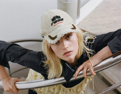 A person with long blonde hair is wearing a distressed beige cap and a black collared shirt. They are holding onto a metal railing, with a curious and expectant expression. The setting suggests an indoor location with light wood flooring and white surroundings, creating a contrast with their attire. The overall composition evokes a sense of casual curiosity.