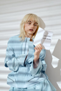 A person with light blonde hair and dark lipstick stands against a textured backdrop, casting soft shadows. They are dressed in a shiny, light blue outfit with billowing sleeves. Their expression is thoughtful, as they hold a crumpled paper, focusing intently on it. The ambiance is modern and artistic, highlighting fashion and introspection.