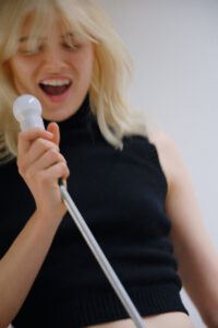 A person with light hair joyfully holds a light bulb attached to a fixture, resembling a microphone. They are dressed in a sleeveless black top and appear to be singing with enthusiasm. The scene gives a sense of casual, playful expression, with the subject exuding an energetic and lively vibe against a soft, neutral background.
