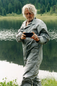 An older individual with white hair and glasses is outdoors, wearing a gray jacket. They are holding a smartphone with both hands, seemingly capturing a moment. In the background, a serene lake reflects the lush green forest surrounding it. The scene suggests a peaceful, natural setting with a focus on technology amidst nature.