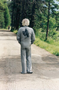 A person with light hair stands in the middle of a dirt path, surrounded by tall, green trees. The individual is wearing a matching set of gray clothes, walking away from the viewer. The scene is tranquil, capturing the essence of a serene, wooded area. Another person is visible in the distance, enhancing the sense of depth and solitude.