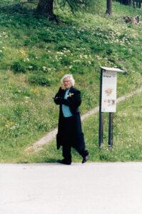 An individual with curly white hair stands on a path, surrounded by a lush, blooming meadow with various wildflowers. Wearing a dark coat and holding a tablet or book, the person appears thoughtful, glancing sideways. A wooden informational sign stands nearby. Trees and green grass fill the background, evoking a serene, natural setting.