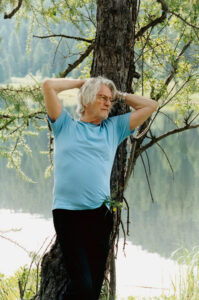 An older man with white hair leans against a tree, arms folded behind his head. He wears a light blue shirt and dark pants. The backdrop features a serene lake and lush, green forest, creating a peaceful atmosphere. Sunlight filters through the leaves, highlighting the tranquil, natural setting. The scene evokes relaxation and contemplation.