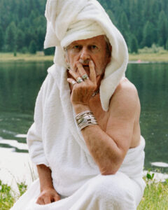 A person sits near a tranquil lake, surrounded by lush greenery. They are dressed in a white robe with a towel wrapped around their head. Adorned with numerous rings and bracelets, they rest their hand near their face in a contemplative manner. The background features a dense forest, enhancing the serene and reflective scene.