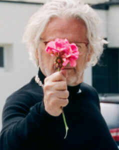 A person with white hair, wearing glasses and a black outfit, holds a vibrant pink flower up to their face. The background is blurred, featuring neutral color tones, adding focus to the individual and the flower. The scene conveys a mix of whimsical and serene elements, with the bright flower contrasting against the dark clothing.