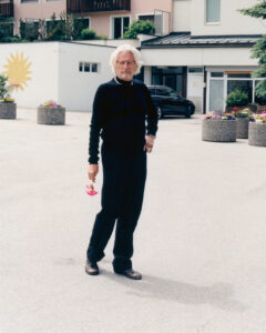 An older man with white hair and glasses stands on a paved area, dressed in black. He holds a small pink object in his hand. Behind him, there are modern buildings adorned with greenery, flower pots, and a parked car. The environment appears calm, with pastel-colored walls and a large cheerful illustration nearby.