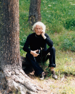 An individual with light hair rests against a tree trunk in a grassy area. Dressed in a black outfit with rolled-up pants, they hold a small device in one hand while leaning their head on the other. The setting is natural, with green grass and some yellow flowers nearby, creating a serene and contemplative atmosphere.