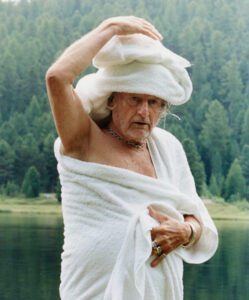 An elderly man, wrapped in a white towel with another on his head, stands by a tranquil body of water. He appears thoughtful, with lush greenery forming a serene backdrop of towering trees. He holds the towel with one hand while raising the other, displaying numerous rings and a bracelet, creating a peaceful and contemplative scene.