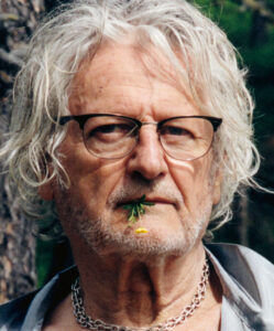 An older person with shaggy white hair and glasses stands outdoors. They have a relaxed beard and are wearing a shiny silver chain necklace. A small yellow flower and green stems are playfully held between their lips. The background consists of blurry green foliage and a tree, suggesting a natural, peaceful setting. The expression conveys wisdom and tranquility.