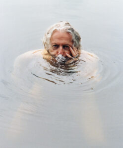 A person with white hair is partially submerged in water, creating ripples around them. They rest their head on one hand, with their face just above the surface, exhaling bubbles. The surrounding water is calm, with a reflective quality that adds a serene, contemplative atmosphere. The gentle play of light creates soft gradients across the scene.
