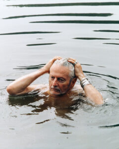 An elderly man stands in the calm waters, with his hands clasping his head, revealing a thoughtful or introspective expression. He wears multiple rings and a bracelet, suggesting a fondness for accessories. The water reflects the play of light on its surface, creating a serene backdrop. The overall mood suggests contemplation or a moment of quiet reflection.