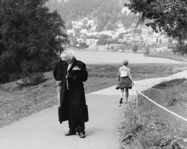A serene pathway winds through a lush landscape, offering a captivating view of distant buildings nestled among hills. Two individuals walk in opposite directions; one clad in a long coat, the other in more casual attire. Tall trees frame the scene, while the gentle curve of the path suggests a tranquil rural or suburban setting.