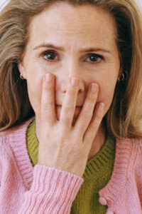 A person with light hair wearing a pink sweater over a green top partially covers their face with their hand, revealing only the upper part of their face and eyes. The fingers, adorned with a subtle sheen, create an intriguing barrier. Small hoop earrings accentuate the ears, adding a touch of elegance to the casual attire.