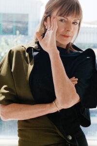 A confident person with light brown hair and bangs gazes directly. One hand touches the side of the face, while the other arm is folded. The person is wearing a stylish, structured garment in olive and black, adorned with a leopard-print button, indoors. Sunlight creates a soft glow in the background, which features a blurred view.