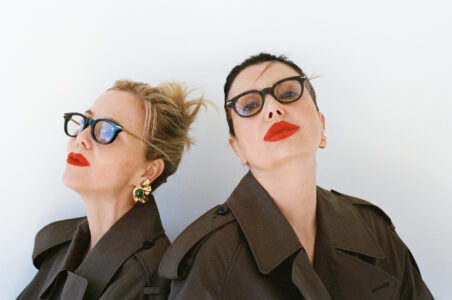 Two individuals stand back-to-back against a plain backdrop, wearing glasses and trench coats. Their expressions are poised, with striking red lipstick. One has blonde hair styled up, adorned with bold green and gold earrings, while the other has dark hair tied back, wearing gold earrings. The overall vibe is stylish, sophisticated, and bold, exuding confidence.