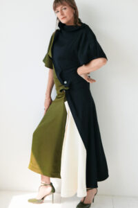 A person stands against a light-colored wall, wearing a stylish, asymmetrical dress with green, blue, and white panels. The dress has a high neckline and a prominent button detail at the waist. They are wearing matching green high heels. Their hair is styled simply, and they have a poised and confident expression.