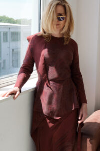 A woman with shoulder-length blonde hair stands by a large window, partially casting a shadow from the sunlight. She wears stylish sunglasses and a unique, intricately patterned, burgundy dress. The background shows a view of a modern building complex surrounded by greenery. The composition creates a contrast between the indoor setting and the outdoor urban landscape.