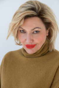 A person with medium-length, light blonde hair and vibrant red lipstick is wearing a high-necked, brown sweater. The expression is focused and confident. The background is plain and light, highlighting the individual’s facial features. The gaze is direct, creating a sense of engagement and presence. Their attire suggests a combination of elegance and warmth.