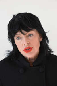 A person with black hair and vivid red lipstick gazes intently, dressed in a black garment with prominent round buttons. The expression is contemplative, set against a neutral background, emphasizing the stark contrast of dark hair and attire with the bright lip color, creating a striking and memorable visage. The overall mood is introspective and poised.