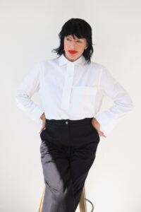 A person with dark hair stands confidently against a plain background, dressed in a crisp white shirt and high-waisted black pants. The posture is relaxed, with hands resting on the hips, and a thoughtful expression on their face. Subtle light shadows accentuate the simplicity and elegance of the overall scene, creating a minimalist aesthetic.