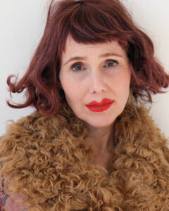 A person gazes directly outward, with short, wavy, reddish-brown hair framing their face. They wear bright red lipstick and have an expressive look. A luxurious, curly, tan-colored textured garment envelops their neck. Soft lighting creates a gentle contrast, highlighting their features against a neutral background, emphasizing a warm, artistic, and somewhat vintage aesthetic and style.