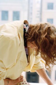 A person with curly auburn hair bends forward indoors, wearing a pale yellow coat over a patterned outfit. The soft lighting casts gentle shadows, hinting at a relaxed atmosphere. The background features a blurred view of tall buildings seen through a window speckled with raindrops, adding a contemplative and serene mood to the scene.