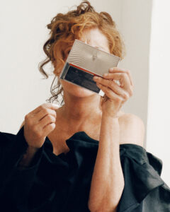 A person with curly, reddish hair is holding a compact mirror, partially covering their face. They are wrapped in a dark, off-the-shoulder garment. The background is a plain, light-colored wall. The overall composition conveys a sense of privacy and introspection, with a subtle play of light and shadow on the person's skin and attire.