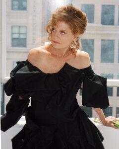 A woman with curly, reddish-brown hair is looking out a window, surrounded by cityscape buildings. She wears an off-the-shoulder black dress with voluminous fabric details. Her expression is contemplative, and natural light enhances her features. The scene conveys a sense of elegance and introspection, blending organic and urban elements against a soft, sunlit backdrop.