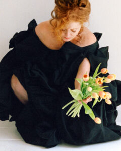 A person with red hair is gently holding a bouquet of orange tulips. They are wearing a voluminous black dress, sitting gracefully on a light-colored floor, creating a striking contrast between the dark fabric and the soft tones of the flowers and surroundings. Their expression is calm and contemplative, adding a serene elegance to the scene.