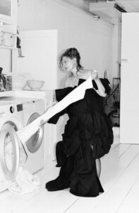 A woman with curly hair, wearing an elegant, voluminous black dress, interacts with a washing machine in a domestic setting. She holds a long, white piece of fabric. The scene includes white-paneled walls, cabinets with stacked bowls, and neatly folded towels. The monochromatic palette gives a classic and stylish atmosphere to the setting.