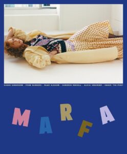 A person with curly hair reclines on the floor in a relaxed pose, dressed in a long, patterned dress and a pale yellow coat. The background is a minimalist, well-lit interior with a large window. The setting and clothing evoke a sense of casual elegance and modern art, with soft colors enhancing the serene atmosphere.