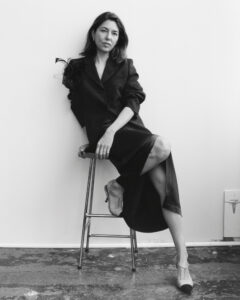 A woman sits on a stool against a plain backdrop. She wears a dark outfit with a floral embellishment on the shoulder. One leg is elegantly crossed over the other, revealing stylish two-tone shoes. Her relaxed posture and contemplative expression convey an air of calm confidence. The scene exudes a minimalist, sophisticated atmosphere.