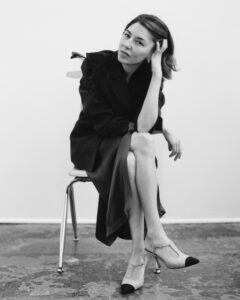 A contemplative individual sits on a simple chair, dressed in a sophisticated outfit with artistic floral details on the shoulder. Their posture is relaxed, with one leg crossed over the other and a hand gently supporting their face. Their gaze is direct and thoughtful. They wear elegant high heels. The setting is minimalist, with a plain background and textured flooring.