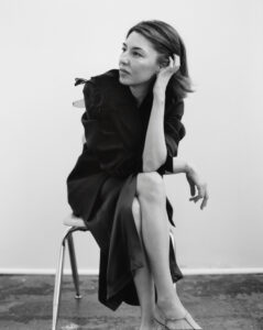 A woman is seated on a simple chair, gazing thoughtfully into the distance. She wears a dark, elegant outfit with a large floral detail on the shoulder. Her posture is relaxed, with a hand resting on her cheek and one leg crossed over the other. The setting is minimalistic, with a plain background and a textured floor.