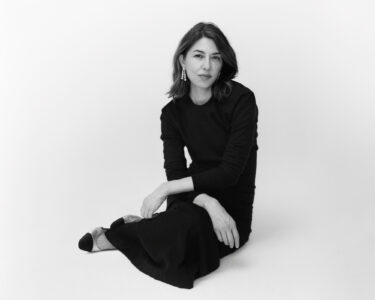 A woman with shoulder-length hair and dangling earrings sits gracefully on a plain surface, dressed in a long, elegant dark dress. Her expression is calm and introspective. She delicately holds her hands together, one resting on her lap. Her poised posture and simple backdrop create a serene, minimalist atmosphere, emphasizing her thoughtful demeanor and understated elegance.