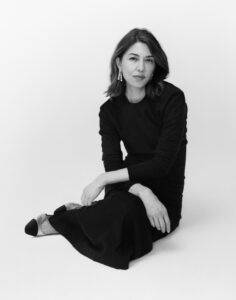 A person with shoulder-length hair sits gracefully on a light surface. They are wearing a long, dark dress and stylish shoes with a contrasting toe. Elegant earrings dangle, adding a touch of sophistication. The setting is simple and minimalist, emphasizing the subject’s poised and contemplative expression against a plain background, creating a sense of calm and elegance.