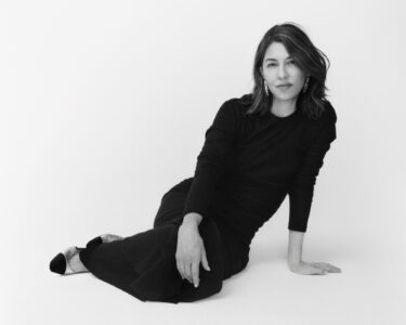 A person with shoulder-length hair, wearing a sleek, long-sleeved black dress, sits gracefully against a plain background. They lounge slightly, one hand supporting their posture on the floor. Subtle earrings add an elegant touch. Their expression is calm and confident. Their legs are elegantly positioned, with shoes featuring contrasting colors, enhancing the overall sophisticated and minimalist aesthetic.