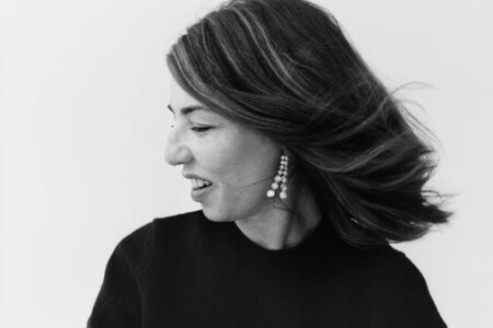 A woman with shoulder-length hair is turned to the side, her head slightly tilted down. The wind appears to flow through her hair, adding dynamic movement. She's wearing a black top and large, dangling earrings with multiple gemstones. Her expression is soft and contemplative, conveying a sense of calm and introspection. The background is plain and light.