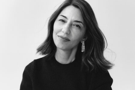 A woman with shoulder-length dark hair is captured in a serene, close-up, black-and-white portrait. She wears a black top and a distinctive dangling earring on one ear. Her expression is calm and thoughtful, softly engaging the viewer. The background is an unobtrusive solid color, drawing focus to her contemplative gaze and elegant appearance.