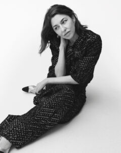 A woman, dressed in an elegant, textured outfit, sits with one leg extended forward. Her head tilts slightly as she rests her chin on her hand, appearing deep in thought. Her gaze is focused in the distance, and her hair falls loosely down, blending with the monochrome setting. The scene conveys a serene, contemplative mood.