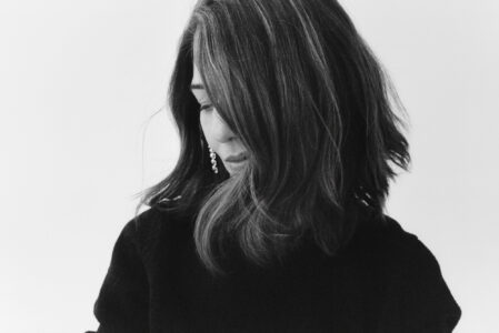 A black and white portrait depicts a person with shoulder-length hair partially covering their face. They are wearing a dark top and have an elegant, dangling earring. The person appears contemplative or introspective, gazing downward with a soft, gentle expression. The overall mood is serene and artistic, capturing a candid moment of reflection.