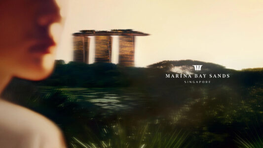 A serene scene features a blurred profile of a person in the foreground, gazing at a striking architectural structure with three illuminated towers and a park-like roof. The background transitions into lush greenery and a water surface, all under a soft, warm sky that creates a tranquil and contemplative atmosphere.