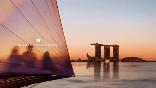 A serene waterfront scene captures the early evening sky painted in soft hues of orange and blue. In the distance, a modern structure with three illuminated towers stands prominently. The foreground features the slightly blurred outline of a sailboat, its sail catching the last light. Reflections shimmer gently on the calm water, evoking a sense of tranquility.