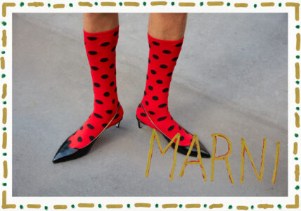Vibrant red socks adorned with bold, black spots extend just below the knee. Paired with sleek, black pointed-toe heels, they create a striking contrast against a light gray floor. The outfit hints at a playful yet chic fashion statement, blending whimsical patterns with elegant footwear. A decorative border frames the scene, adding an artistic touch.
