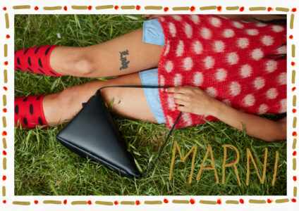 A person lies on grass, wearing a red garment with white spots. Their legs are adorned with red socks featuring black spots, and a tattoo is visible on one thigh. A black triangular bag rests across their lap. A decorative border surrounds the scene, adding an artistic touch to the composition.