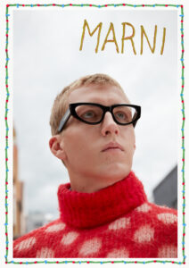 A person gazes upwards with a contemplative expression, wearing bold, black-framed glasses. The striking red turtleneck features a large polka dot pattern in white. The background shows an urban setting with a soft focus on buildings and a cloudy sky. The scene conveys a modern, artistic atmosphere with an emphasis on fashion and individuality.