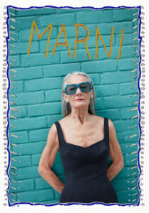 A confident elderly woman stands against a teal brick wall. She wears oversized, stylish blue sunglasses and a sleek black sleeveless dress. Her long gray hair flows naturally, adding to her dignified presence. The frame has a whimsical border with doodles, highlighting her vibrant and bold personality while showcasing a modern twist on elegance and style.