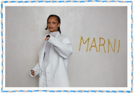 A person is standing against a plain background, wearing a stylish white outfit with an oversized jacket. The individual has a confident posture and braided hairstyle. The border is decorated with a blue and orange pattern. The overall atmosphere exudes a sense of fashion-forward elegance and contemporary style, highlighting the sophistication of the attire.